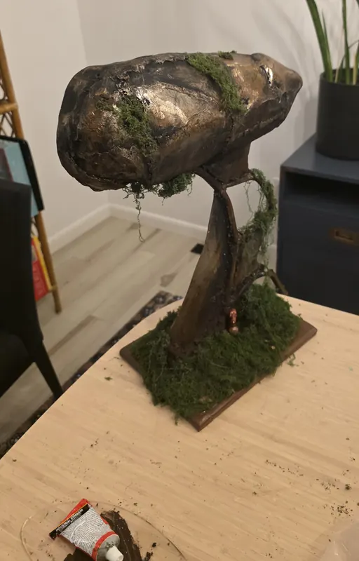 Mixed media sculpture of a bronze, moss-covered mass on a twisted trunk rising from a mossy base
